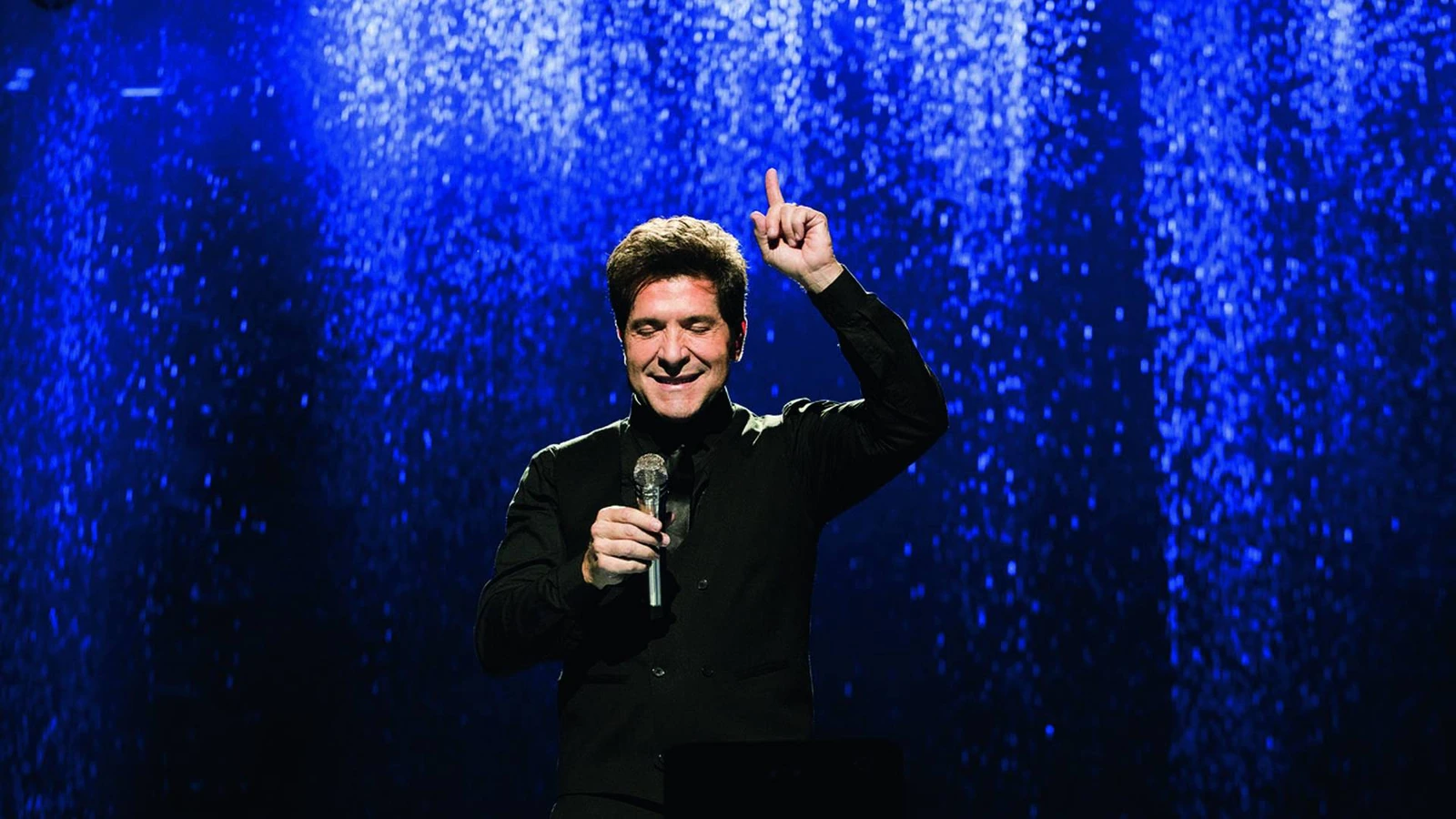 Daniel canta com o microfone na mão e aponta para o alto, diante de um painel azul.