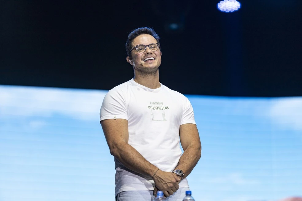 Deive Leonardo em plano médio, com camiseta branca e microfone de cabeça, diante do painel azul.