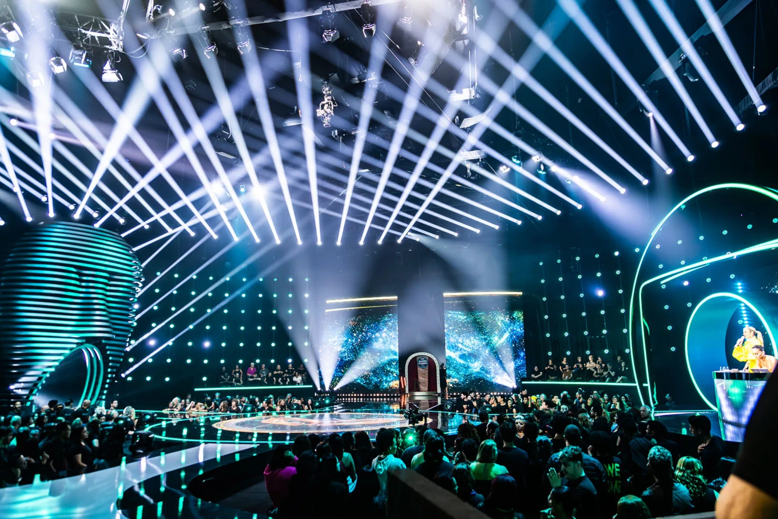A wide view of The Masked Singer Brasil studio, with the audience and beams of stage light.